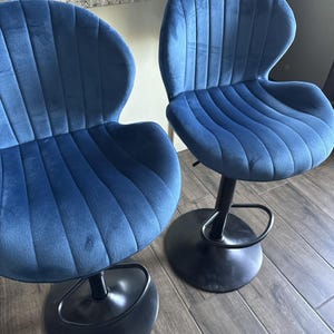 May include: Two bar stools featuring deep blue velvet upholstery with vertical channel tufting. Each stool has a curved backrest, a contoured seat, and a black metal pedestal base with a circular footrest and a round floor plate for stability.