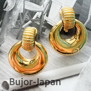 May include: A pair of gold-toned earrings with a large, circular design. The earrings have a polished, reflective surface and a textured, ribbed detail at the top. The text "Bujor-Japan" is visible in the image.