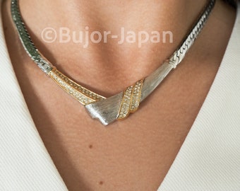 May include: A silver and gold necklace with a V-shaped design and clear stones. The necklace is on a model wearing a white shirt.