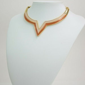 May include: A gold-toned collar necklace with a geometric design. The necklace features a V-shaped pendant with layers of peach and light pink enamel. The necklace is displayed on a white mannequin bust.
