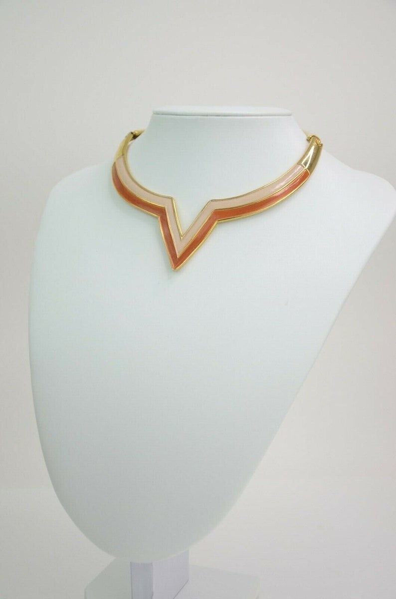 May include: A gold-toned collar necklace with a geometric design. The necklace features a V-shaped pendant with layers of peach and light pink enamel. The necklace is displayed on a white mannequin bust.