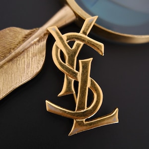May include: A gold-toned metal brooch featuring the interlocking YSL logo. The brooch is positioned next to a gold-toned magnifying glass with a textured handle shaped like a feather, showcasing a polished finish and sharp, defined edges.