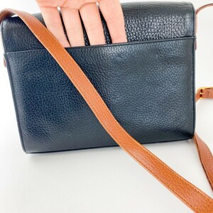 May include: Black leather crossbody bag with a brown strap. The bag has a flap closure and a pocket on the back.