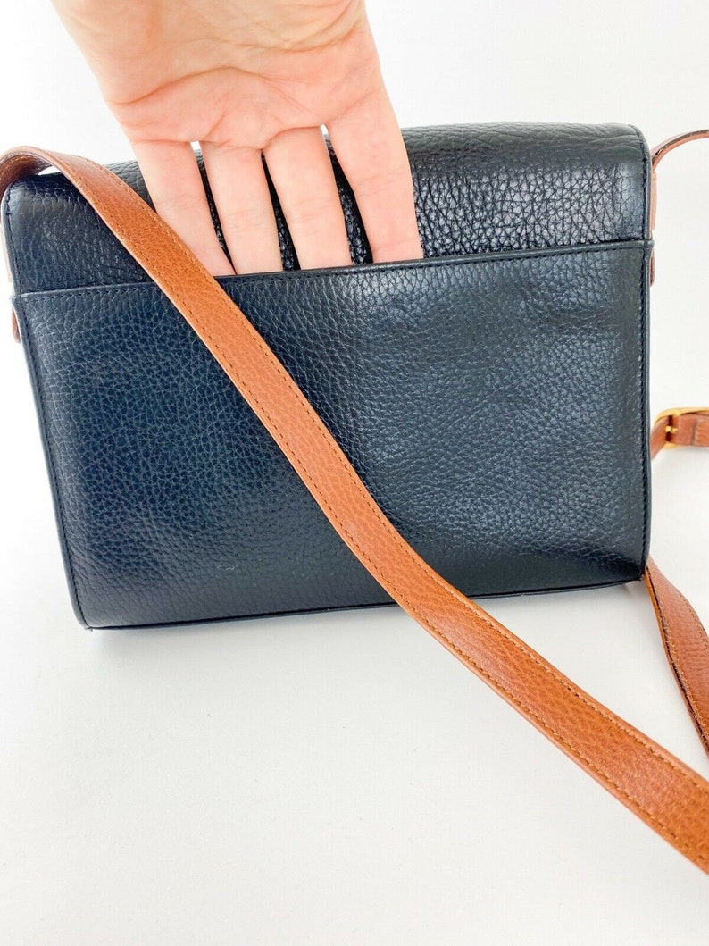 May include: Black leather crossbody bag with a brown strap. The bag has a flap closure and a pocket on the back.