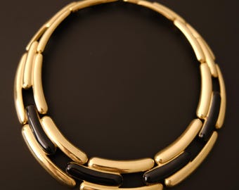 Puede incluir: Un collar circular tipo gargantilla que presenta eslabones de metal dorado pulido y segmentos de resina negra brillante. El diseño consta de dos filas concéntricas de barras curvas y alargadas conectadas para formar una estructura rígida.