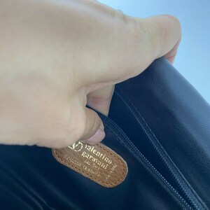 May include: Close-up of a brown leather tag with the text "Valentino Garavani" and "Genuine Leather" in gold lettering. The tag is attached to a black leather bag.