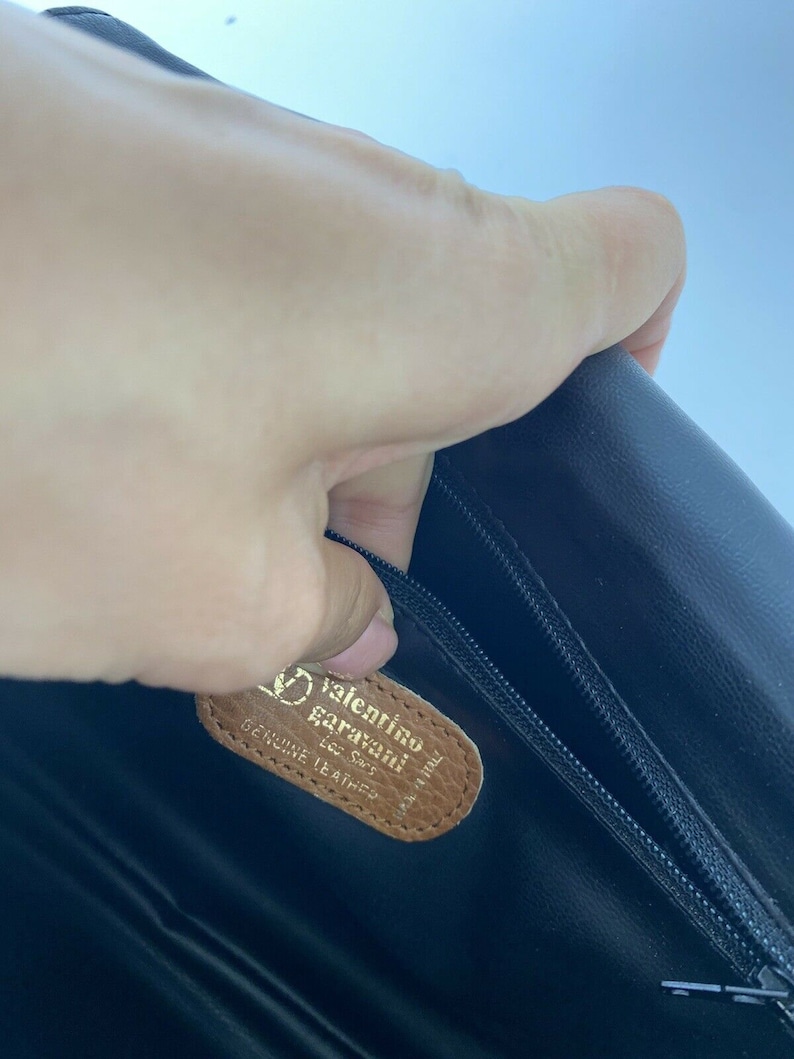 May include: Close-up of a brown leather tag with the text "Valentino Garavani" and "Genuine Leather" in gold lettering. The tag is attached to a black leather bag.