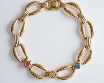May include: Gold-toned chain link necklace with oval links and a clasp closure. The necklace features three colorful gemstone accents: a pink teardrop, a round yellow-green stone, and a square blue stone. The jewelry is displayed on a white background.