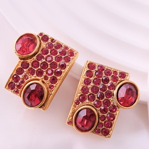 May include: A pair of gold-toned earrings. Each earring features a rectangular shape with a border of small, round, red gemstones. Two larger, oval-shaped red gemstones are set on each earring, one at the top and one at the bottom.