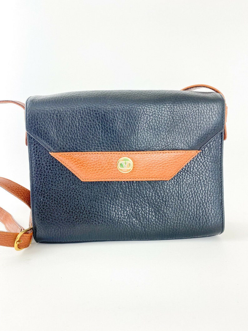May include: A black leather shoulder bag with a brown leather flap and a gold metal clasp. The bag has a textured surface and a long adjustable strap.