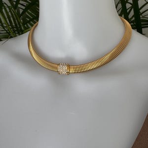May include: A gold-toned choker necklace with a ribbed design. The necklace features a square, central clasp adorned with small, sparkling crystals. The jewelry is displayed on a white mannequin, showcasing its elegant design.