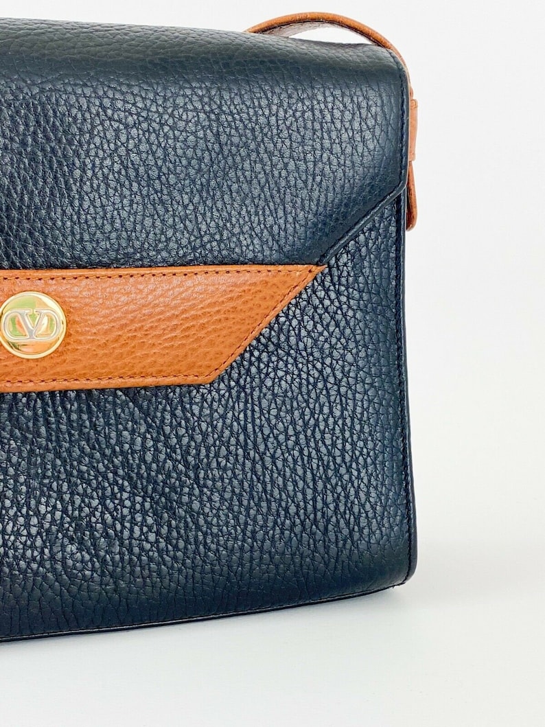 May include: A black leather shoulder bag with a brown leather flap and a gold metal logo. The bag has a textured finish.