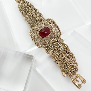 Puede incluir: Un brazalete de cadena dorada con una piedra preciosa roja engastada en un diseño de filigrana dorada.