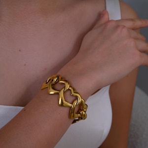 May include: A gold-colored bracelet featuring heart-shaped links. The bracelet is worn on a wrist, with the links forming a chain-like design. The jewelry is a bright, shiny gold color.