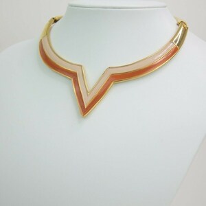 May include: A gold-toned necklace with a V-shaped pendant. The pendant features a layered design with shades of peach and light pink. The necklace is displayed on a white mannequin, showcasing its design.