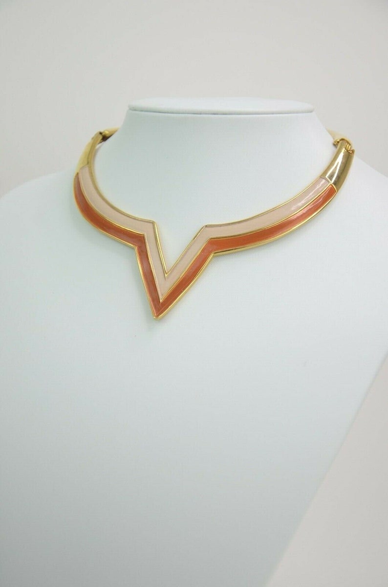 May include: A gold-toned necklace with a V-shaped pendant. The pendant features a layered design with shades of peach and light pink. The necklace is displayed on a white mannequin, showcasing its design.