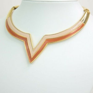 May include: Gold-tone necklace with a geometric V-shaped pendant. The pendant features a layered design with shades of peach and pink enamel. The necklace is displayed on a white mannequin bust, showcasing its design and color.