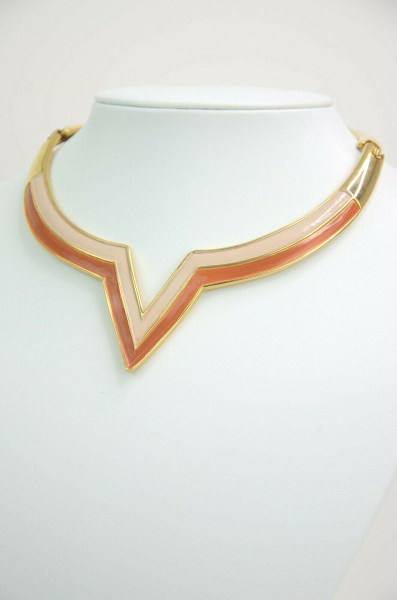 May include: Gold-tone necklace with a geometric V-shaped pendant. The pendant features a layered design with shades of peach and pink enamel. The necklace is displayed on a white mannequin bust, showcasing its design and color.