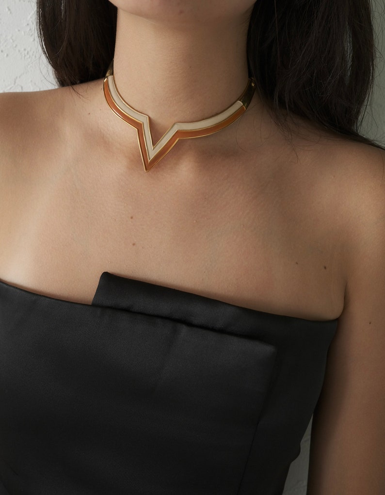 May include: A gold-toned necklace with a V-shaped pendant. The pendant features a geometric design with alternating bands of white and orange enamel. The necklace is worn against a black strapless top.