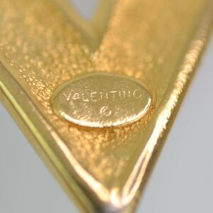 May include: Close-up of a gold-toned metal brooch in a V-shape. The brooch has a textured surface and a small, oval-shaped inset with the word "Valentino" engraved. The overall design is simple and elegant.