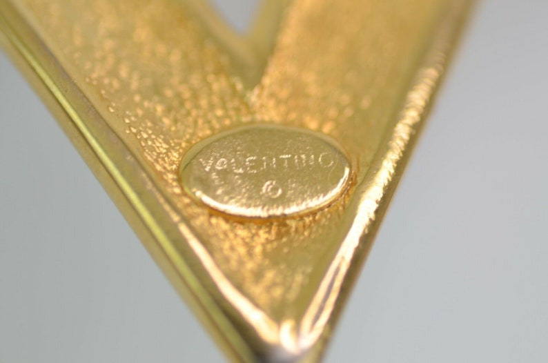 May include: Close-up of a gold-toned metal brooch in a V-shape. The brooch has a textured surface and a small, oval-shaped inset with the word "Valentino" engraved. The overall design is simple and elegant.