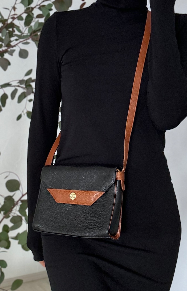 May include: A black leather crossbody bag with a brown leather strap and flap. The bag has a gold-toned button closure.