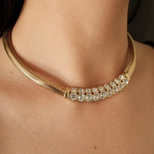 May include: A gold-toned necklace with a mesh chain and a central bar adorned with multiple clear, round gemstones. The necklace is worn around the neck, showcasing its design and sparkle. The gemstones are arranged in two rows.