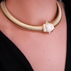 May include: Gold-tone collar necklace with a ribbed design. The necklace features a central rectangular pendant with a white stone and two small, clear accent stones. The jewelry is worn against a black garment.