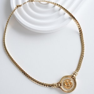 May include: A gold-toned necklace with a circular pendant. The pendant features a double-C logo surrounded by small, clear stones. The necklace has a flat, woven chain design. The jewelry is displayed on a white surface.
