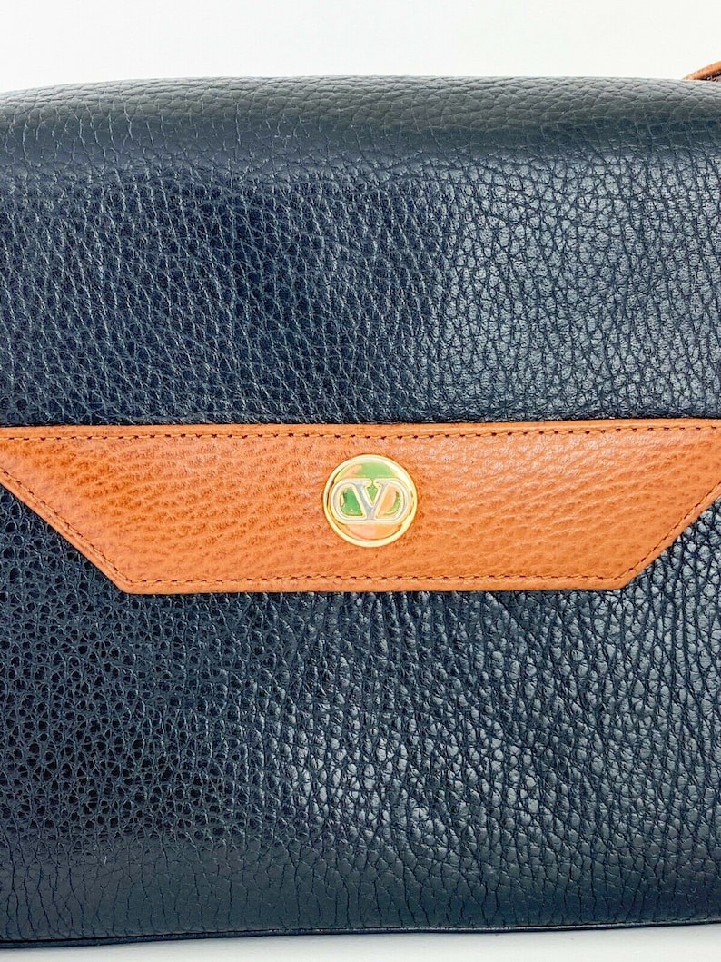 May include: A black leather handbag with a brown leather flap and a gold-toned logo. The bag has a textured surface.