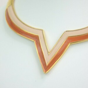 May include: A gold-toned necklace with a geometric V-shaped pendant. The pendant features layers of peach and light pink enamel, outlined in gold. The necklace is displayed on a white surface.