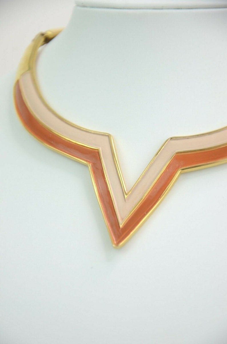 May include: A gold-toned necklace with a geometric V-shaped pendant. The pendant features layers of peach and light pink enamel, outlined in gold. The necklace is displayed on a white surface.