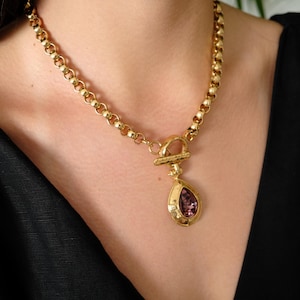 Puede incluir: Collar dorado con un gran colgante en forma de pera con una piedra preciosa púrpura. La cadena es una serie de eslabones redondeados. El collar se lleva sobre una blusa negra.