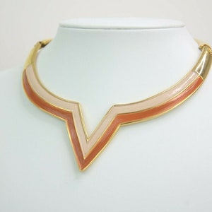 May include: A gold-toned necklace with a V-shaped pendant. The pendant features a layered design with shades of peach and cream enamel. The necklace is displayed on a white mannequin, showcasing its geometric and retro style.