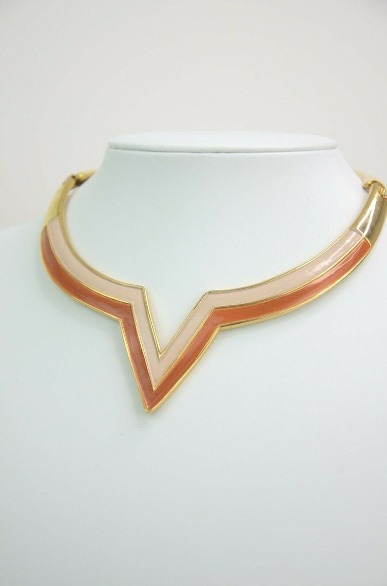 May include: A gold-toned necklace with a V-shaped pendant. The pendant features a layered design with shades of peach and cream enamel. The necklace is displayed on a white mannequin, showcasing its geometric and retro style.