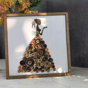 May include: A square framed wall art piece featuring a silhouette of a person with an updo hairstyle. The person's dress is constructed from a dense arrangement of various round buttons in shades of brown, black, tan, orange, and green.