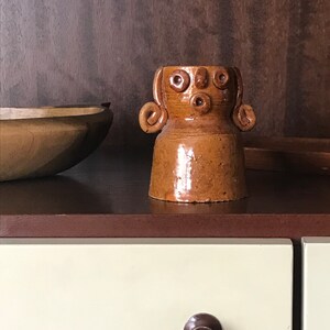 May include: A brown ceramic vase with a face and curly hair. The vase has a textured surface and is sitting on a wooden surface.