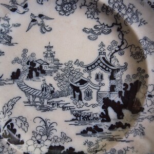 K&ouml;nnte beinhalten: Ein wei&szlig;er Keramikteller mit einem schwarzen, asiatisch inspirierten Landschaftsdesign. Das Design umfasst Pagoden, Br&uuml;cken, V&ouml;gel und florale Elemente. Der Teller hat einen gewellten Rand.