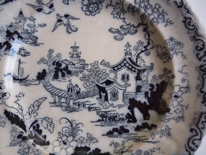 K&ouml;nnte beinhalten: Ein wei&szlig;er Keramikteller mit einem schwarzen, asiatisch inspirierten Landschaftsdesign. Das Design umfasst Pagoden, Br&uuml;cken, V&ouml;gel und florale Elemente. Der Teller hat einen gewellten Rand.