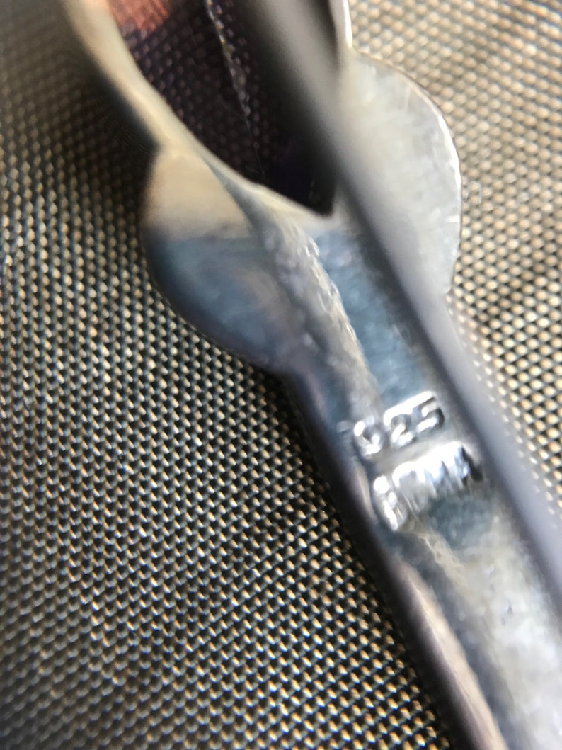 K&ouml;nnte beinhalten: Nahaufnahme eines silberfarbenen Metallobjekts mit dem Text "925" und "1MM" eingepr&auml;gt. Das Objekt hat eine gebogene Form und scheint ein Schmuckbestandteil zu sein. Der Hintergrund ist eine strukturierte, dunkle Oberfl&auml;che.