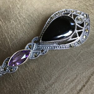 K&ouml;nnte beinhalten: Silberfarbene Brosche mit einem tropfenf&ouml;rmigen schwarzen Onyxstein und einem kleineren, ovalen Amethyststein. Die Brosche hat ein dekoratives Filigran-Design mit kleinen, runden, silberfarbenen Akzenten.