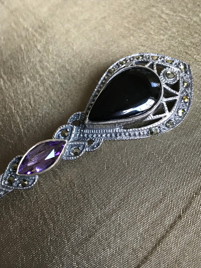 K&ouml;nnte beinhalten: Silberfarbene Brosche mit einem tropfenf&ouml;rmigen schwarzen Onyxstein und einem kleineren, ovalen Amethyststein. Die Brosche hat ein dekoratives Filigran-Design mit kleinen, runden, silberfarbenen Akzenten.