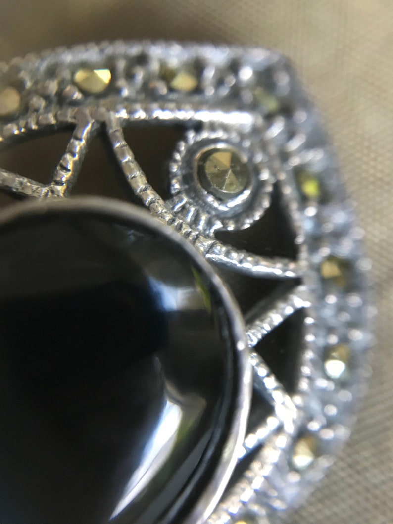 K&ouml;nnte beinhalten: Nahaufnahme einer silberfarbenen Brosche mit einem gro&szlig;en, ovalen schwarzen Stein in der Mitte. Die Brosche hat aufwendige Filigranarbeiten und kleine, goldfarbene Akzente am Rand. Das Design ist aufwendig und von Vintage inspiriert.