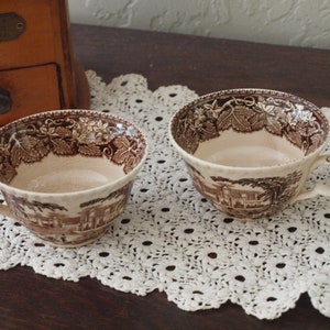 May include: Two vintage teacups in brown and cream. The cups have a detailed landscape design and a leaf pattern around the rim. They are set on a white lace doily. The cups are ideal for tea.