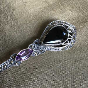 K&ouml;nnte beinhalten: Silberfarbene Brosche mit einem tropfenf&ouml;rmigen schwarzen Stein und einem kleineren, oval geschliffenen lila Stein. Die Brosche ist mit kleinen, runden, silberfarbenen Akzenten verziert. Das Design ist aufwendig und detailliert, mit Vintage-&Auml;sthetik.