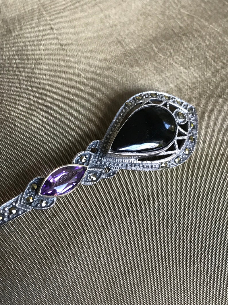K&ouml;nnte beinhalten: Silberfarbene Brosche mit einem tropfenf&ouml;rmigen schwarzen Stein und einem kleineren, oval geschliffenen lila Stein. Die Brosche ist mit kleinen, runden, silberfarbenen Akzenten verziert. Das Design ist aufwendig und detailliert, mit Vintage-&Auml;sthetik.