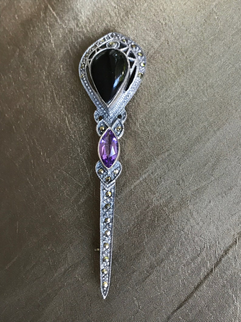 K&ouml;nnte beinhalten: Vintage Brosche in Silberoptik mit einem schwarzen Onyxstein und einem tropfenf&ouml;rmigen Amethysten. Die Brosche ist mit Markasitsteinen besetzt und hat eine lange, spitze Nadel.