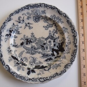 K&ouml;nnte beinhalten: Eine Vintage-Porzellanteller mit blauem und wei&szlig;em Chinoiserie-Design. Der Teller zeigt eine detaillierte Landschaftsszene mit Pagoden, V&ouml;geln und floralen Mustern. Der Teller hat einen gewellten Rand und misst etwa 25 cm im Durchmesser.