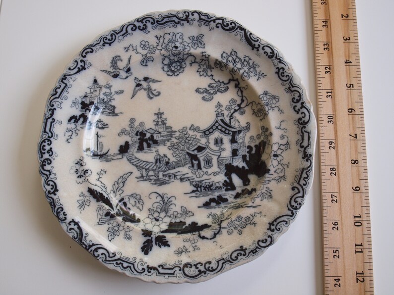 K&ouml;nnte beinhalten: Eine Vintage-Porzellanteller mit blauem und wei&szlig;em Chinoiserie-Design. Der Teller zeigt eine detaillierte Landschaftsszene mit Pagoden, V&ouml;geln und floralen Mustern. Der Teller hat einen gewellten Rand und misst etwa 25 cm im Durchmesser.