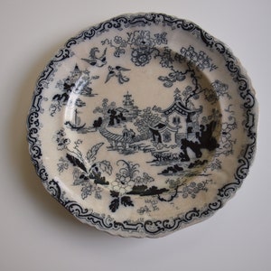 K&ouml;nnte beinhalten: Ein cremefarbener Teller mit einem blau-wei&szlig;en, asiatisch inspirierten Design. Der Teller zeigt eine detaillierte Landschaftsszene mit Pagoden, Br&uuml;cken und V&ouml;geln, umrahmt von einem dekorativen Rand. Ein dekorativer Teller im Vintage-Stil.
